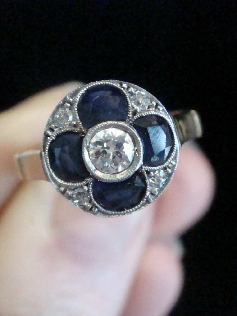 An Art Deco ring featuring a central round diamond surrounded by four dark blue sapphires arranged in a four-leaf clover motif with millegrain platinum borders and a gold shank. View V