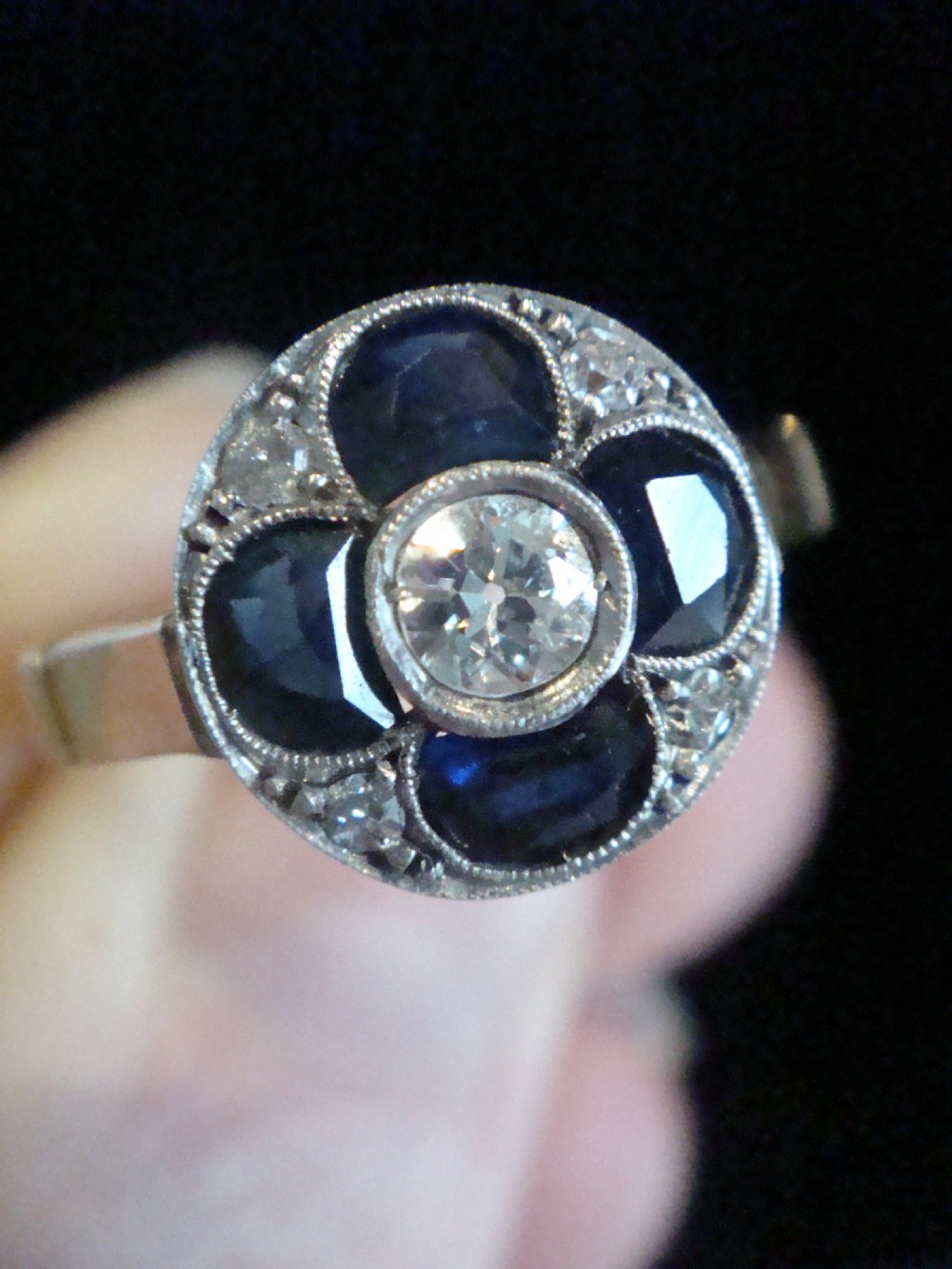 An Art Deco ring featuring a central round diamond surrounded by four dark blue sapphires arranged in a four-leaf clover motif with millegrain platinum borders and a gold shank. View VI