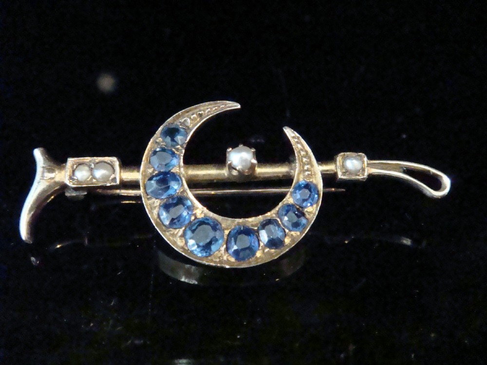 An Edwardian 9ct yellow gold bar brooch featuring a crescent moon motif set with blue sapphires and accented with seed pearls.