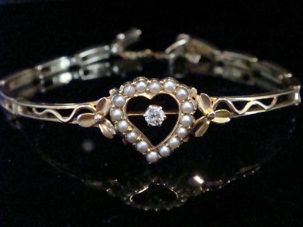 An Edwardian 9ct yellow gold bracelet featuring a central openwork heart set with seed pearls and a single diamond, flanked by sculpted foliage and wavy shoulders.