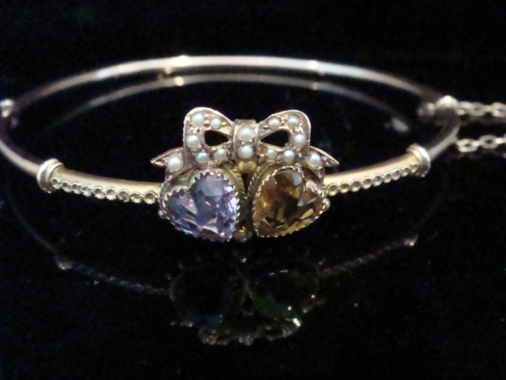 A gold hinged bangle featuring two heart-cut gemstones in purple amethyst and yellow citrine, surmounted by a bow set with small seed pearls.