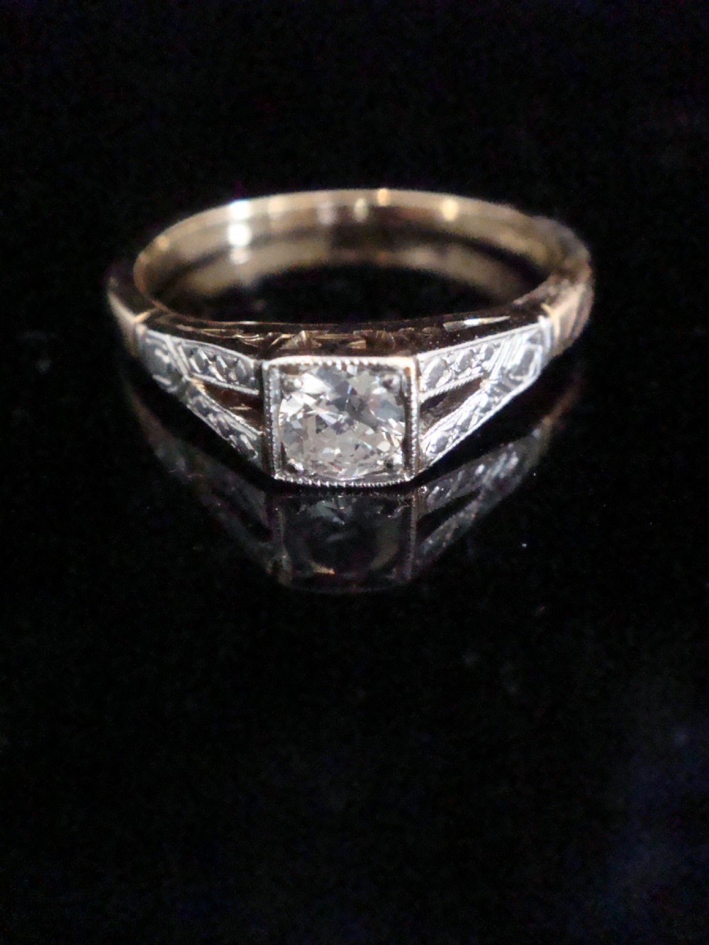 An Edwardian solitaire ring crafted in platinum and gold featuring a central old cut diamond within a square millegrain border and decorated shoulders.