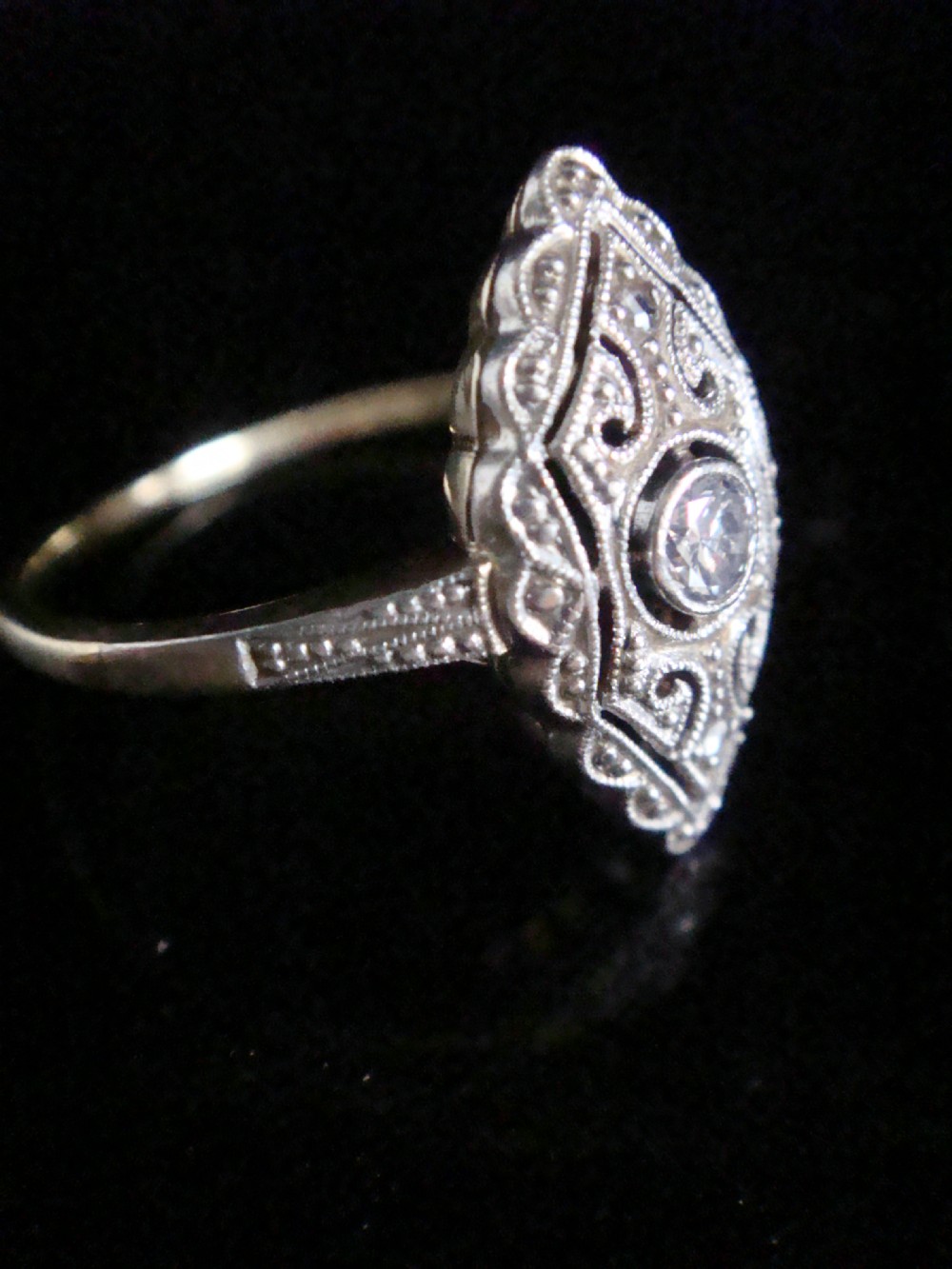 An Art Deco diamond ring featuring an elongated geometric navette setting with intricate filigree work, milgrain detailing, and a central transitional cut diamond. View III