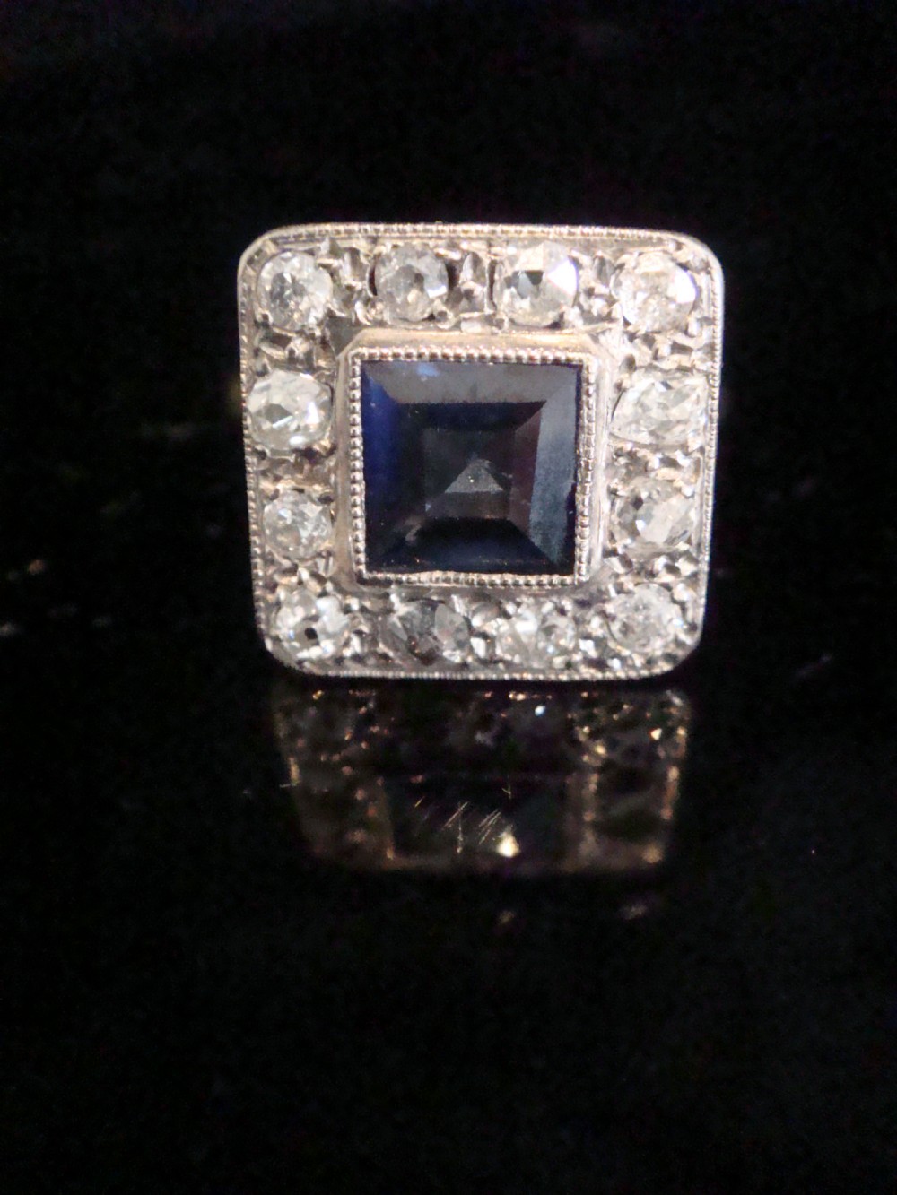 An Edwardian platinum and gold square cluster ring featuring a blue synthetic sapphire surrounded by old cut diamonds with a millegrain edge.