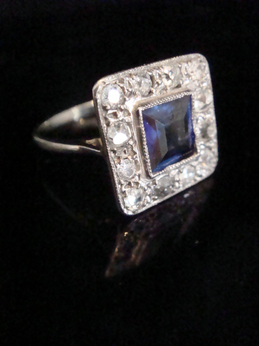 An Edwardian platinum and gold square cluster ring featuring a blue synthetic sapphire surrounded by old cut diamonds with a millegrain edge. View II