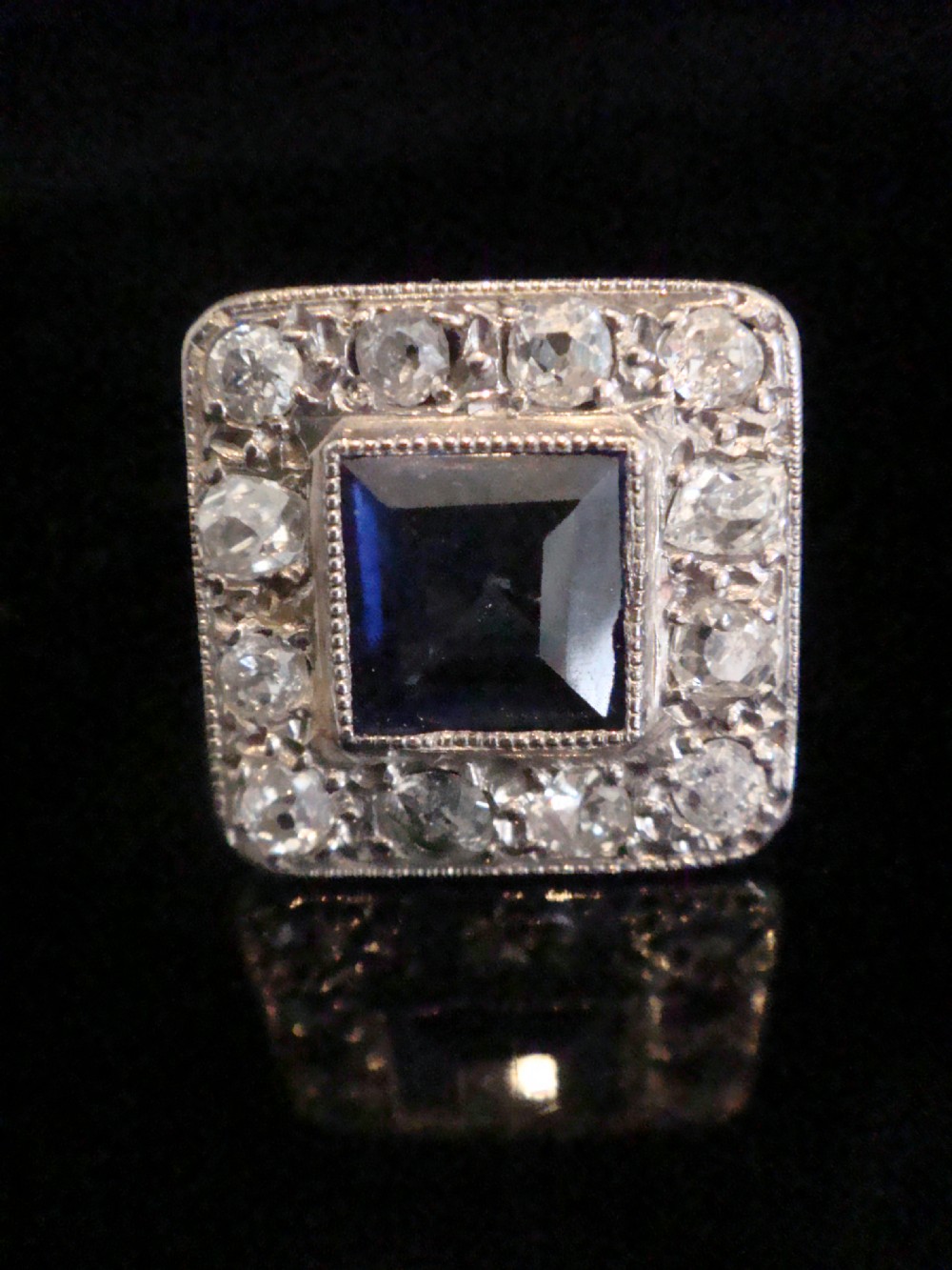 An Edwardian platinum and gold square cluster ring featuring a blue synthetic sapphire surrounded by old cut diamonds with a millegrain edge. View IV