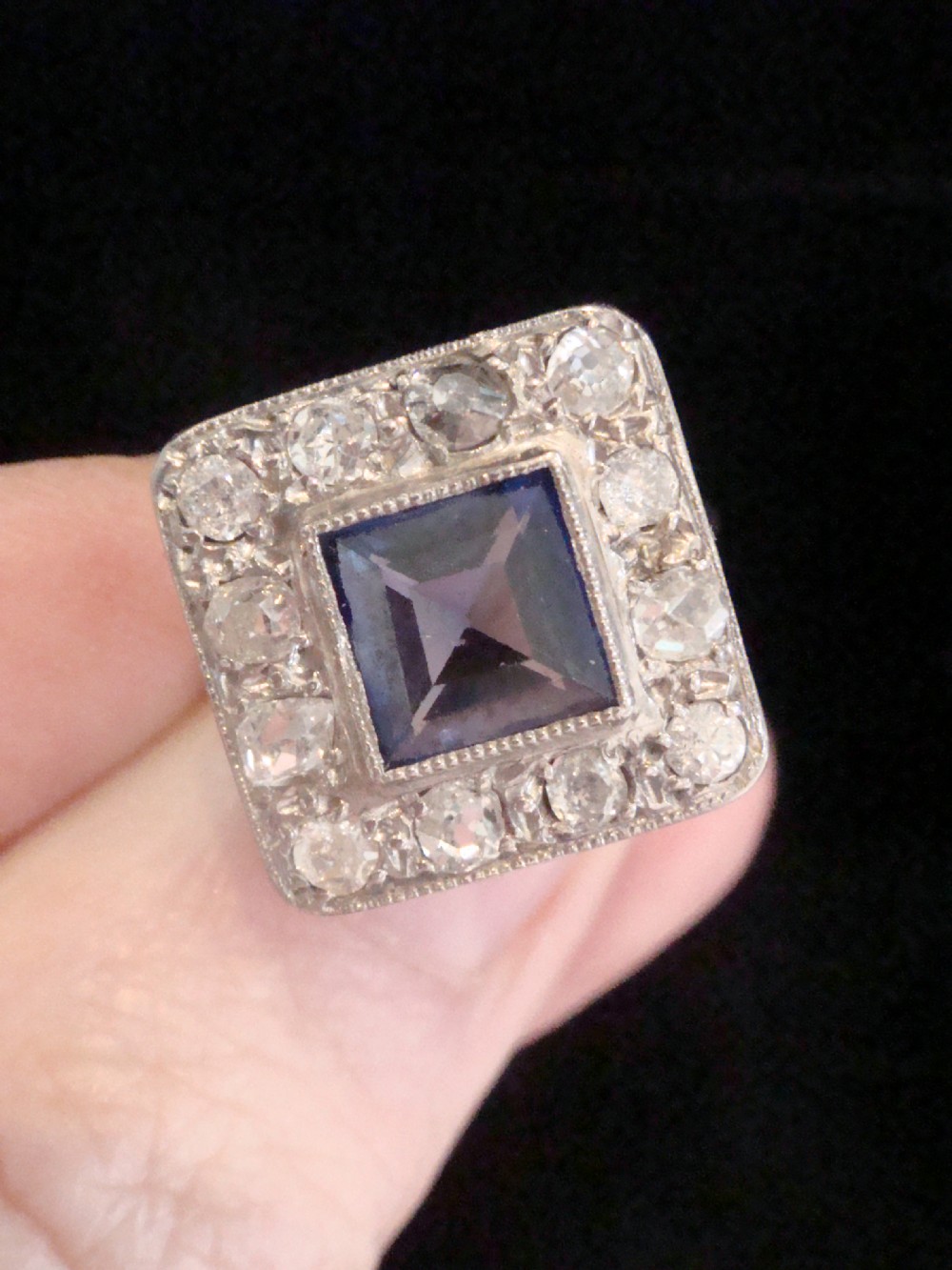 An Edwardian platinum and gold square cluster ring featuring a blue synthetic sapphire surrounded by old cut diamonds with a millegrain edge. View VI