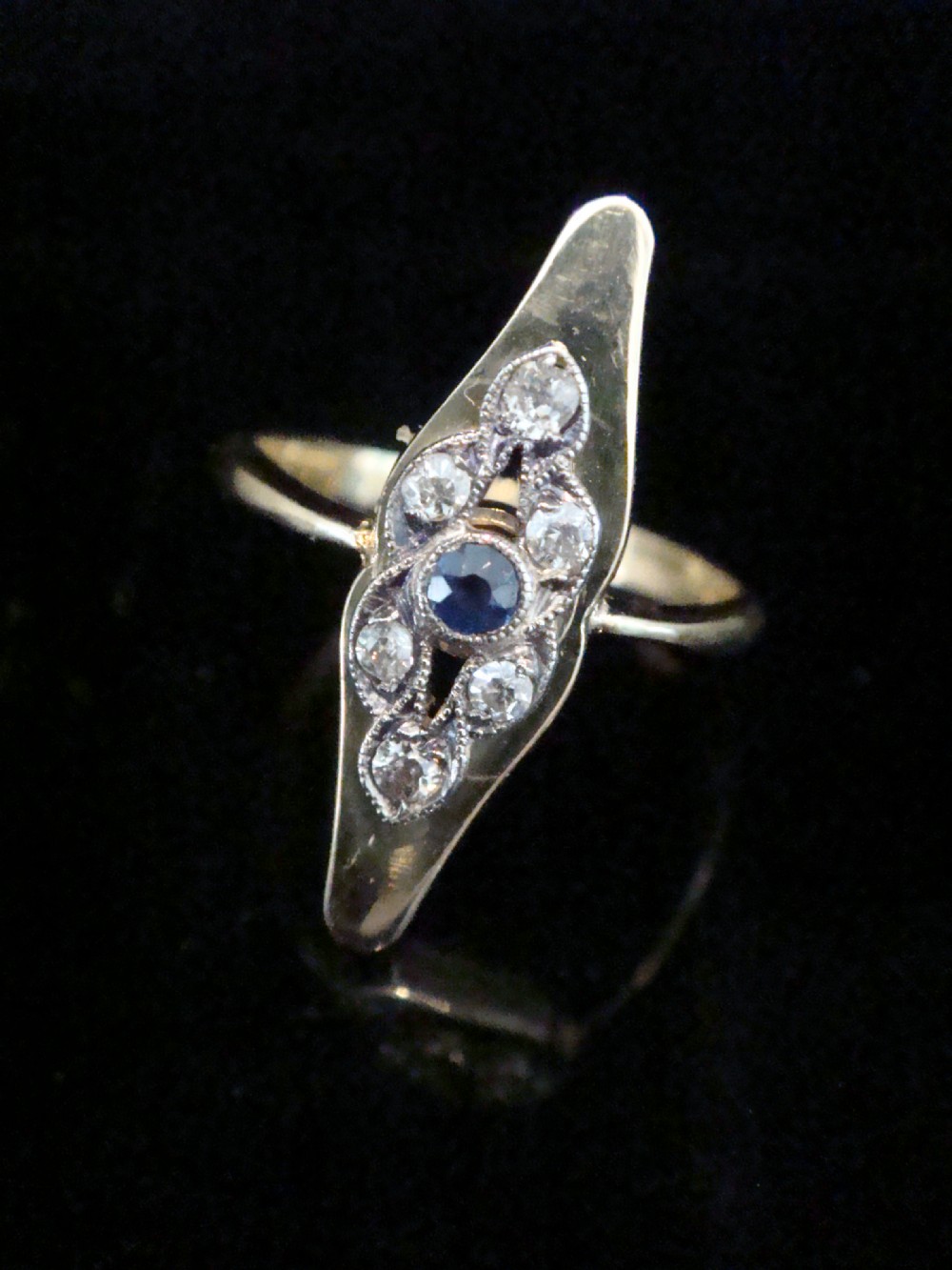 An Edwardian marquise-shaped ring featuring a central round blue sapphire encircled by old cut diamonds set in platinum atop an 18ct yellow gold band.