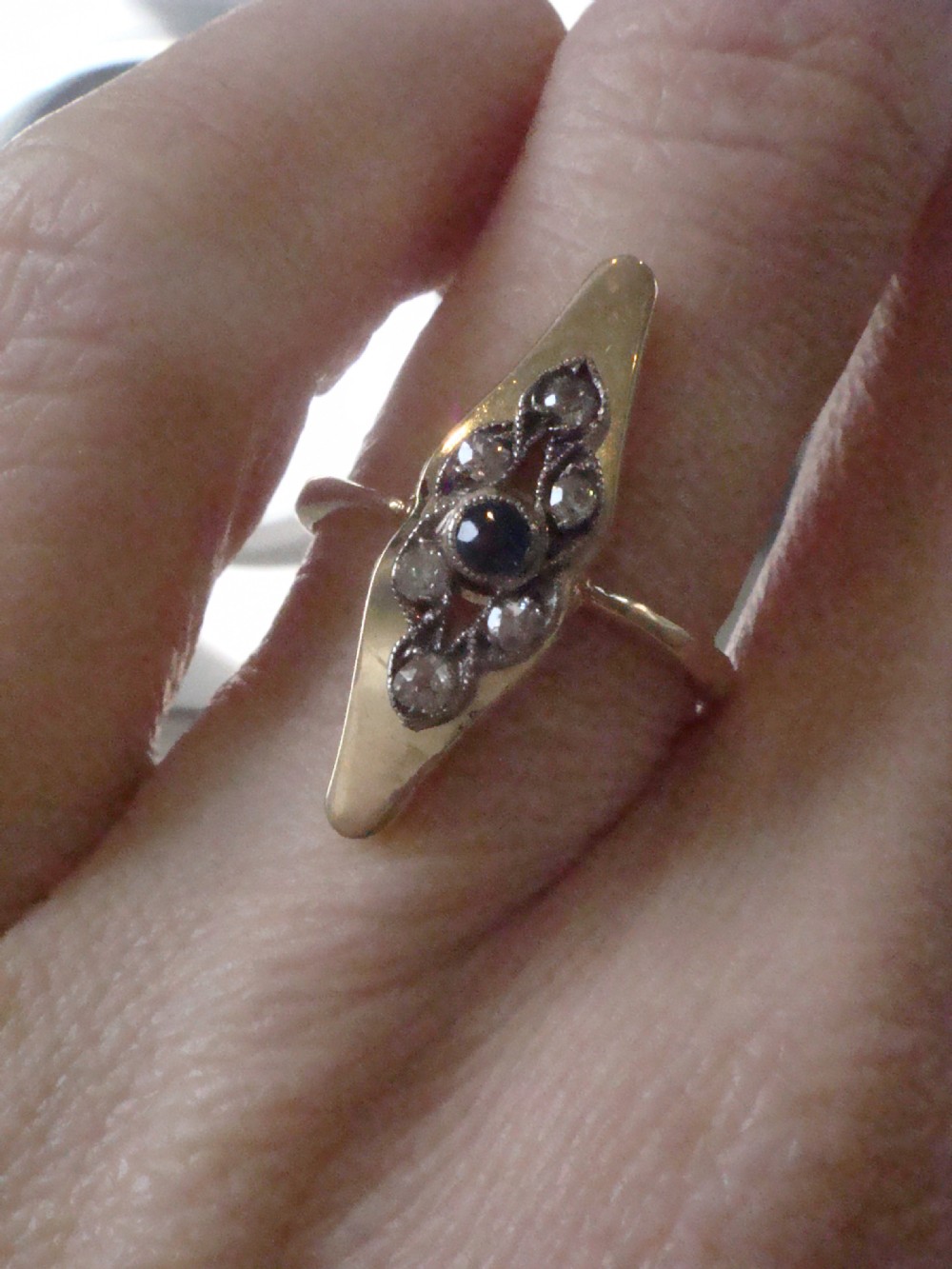 An Edwardian marquise-shaped ring featuring a central round blue sapphire encircled by old cut diamonds set in platinum atop an 18ct yellow gold band. View IV
