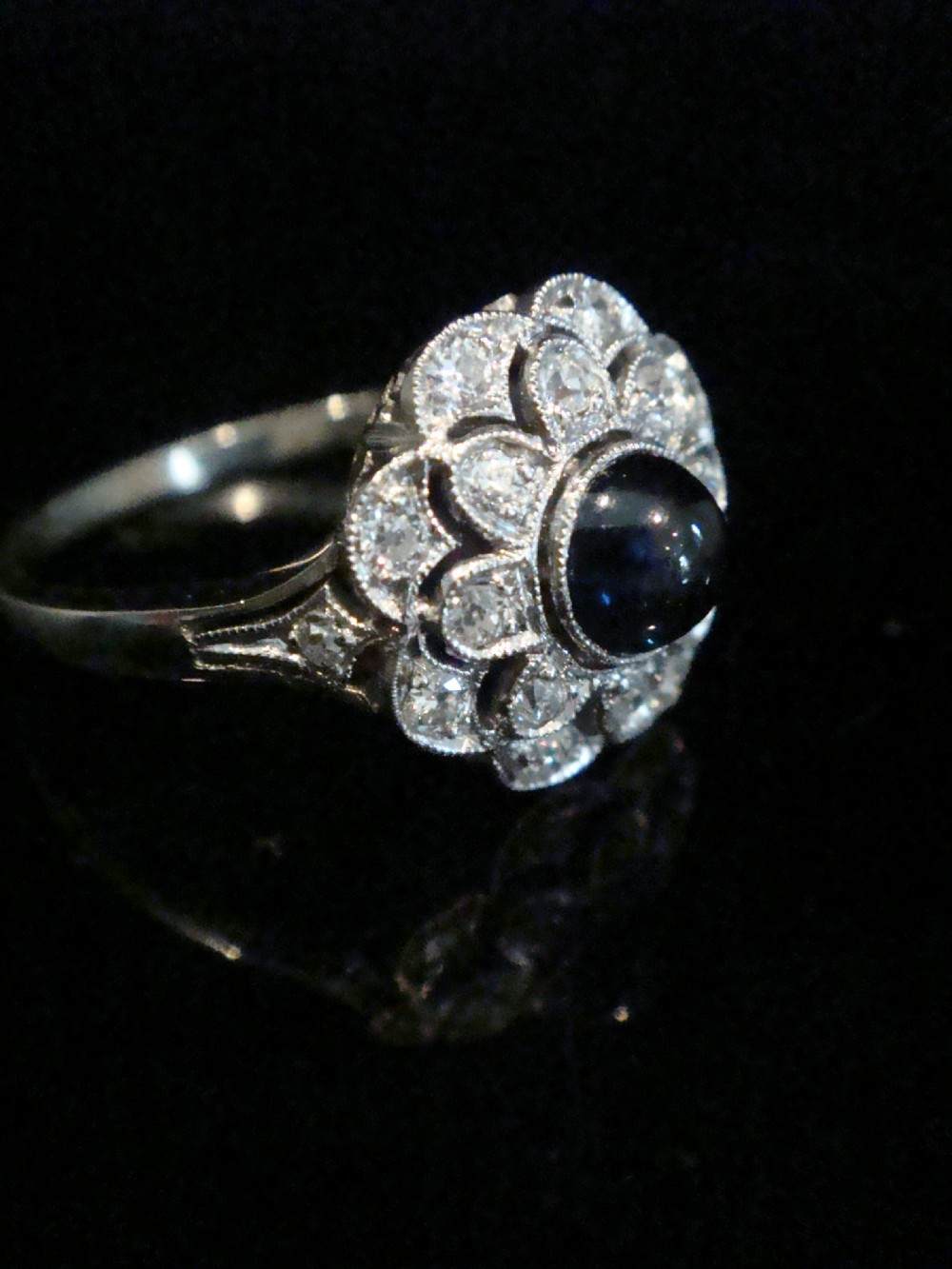 An Art Deco 18ct white gold cluster ring with a central dark blue cabochon sapphire surrounded by double scalloped halos of old cut diamonds.