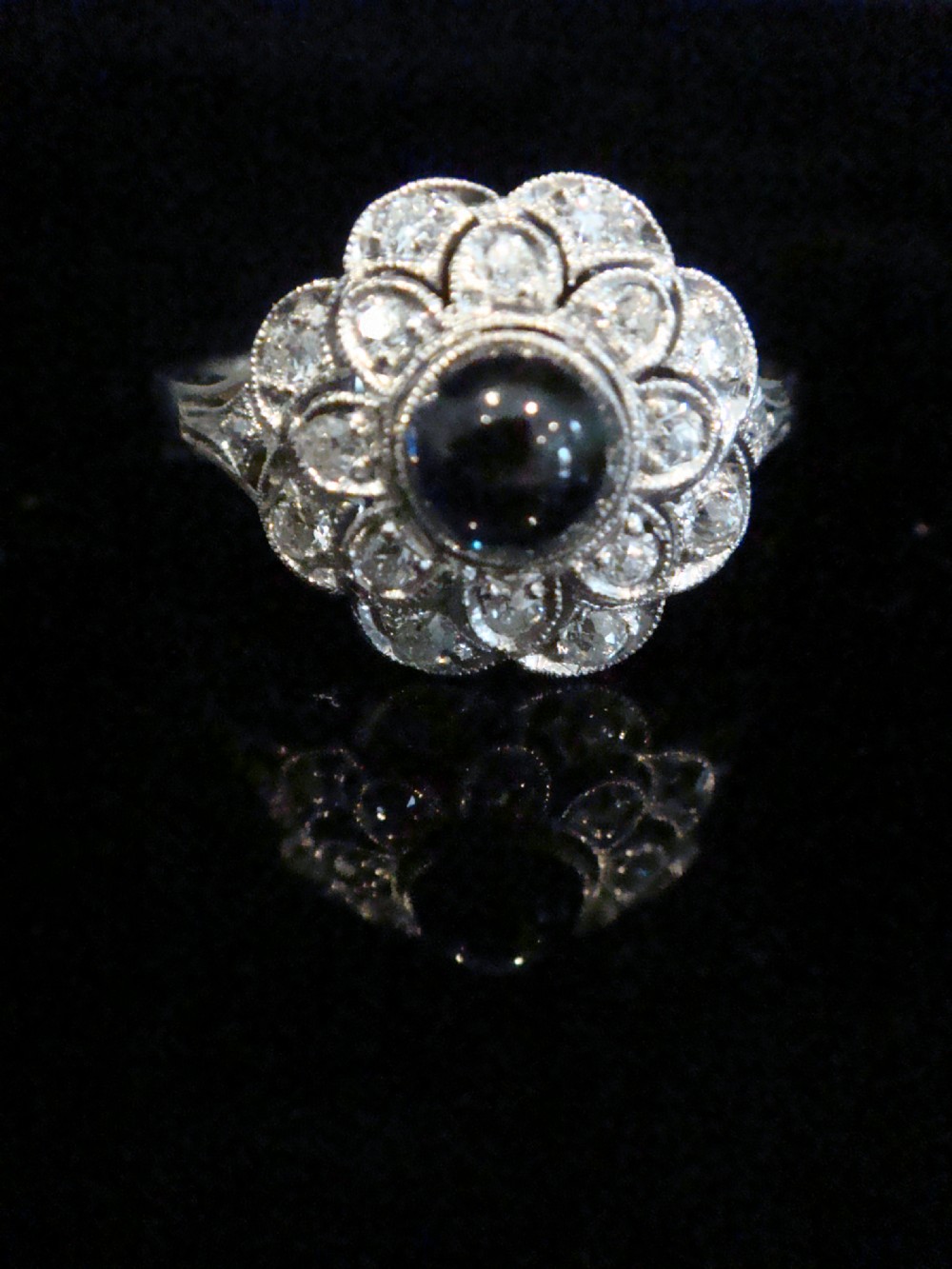 An Art Deco 18ct white gold cluster ring with a central dark blue cabochon sapphire surrounded by double scalloped halos of old cut diamonds. View II