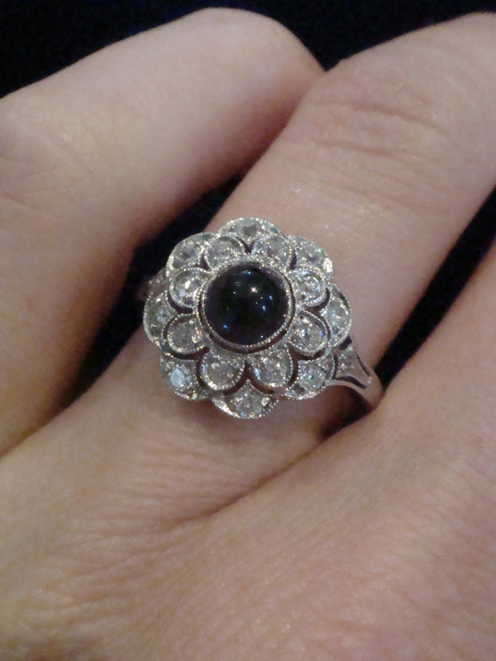 An Art Deco 18ct white gold cluster ring with a central dark blue cabochon sapphire surrounded by double scalloped halos of old cut diamonds. View III