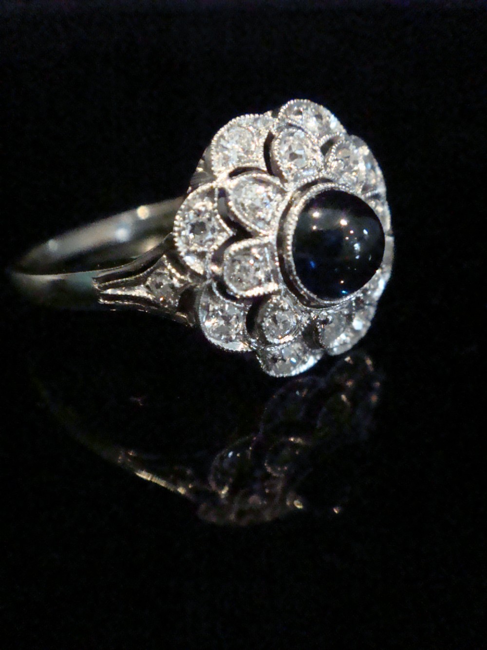 An Art Deco 18ct white gold cluster ring with a central dark blue cabochon sapphire surrounded by double scalloped halos of old cut diamonds. View IV