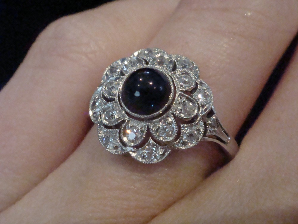 An Art Deco 18ct white gold cluster ring with a central dark blue cabochon sapphire surrounded by double scalloped halos of old cut diamonds. View V