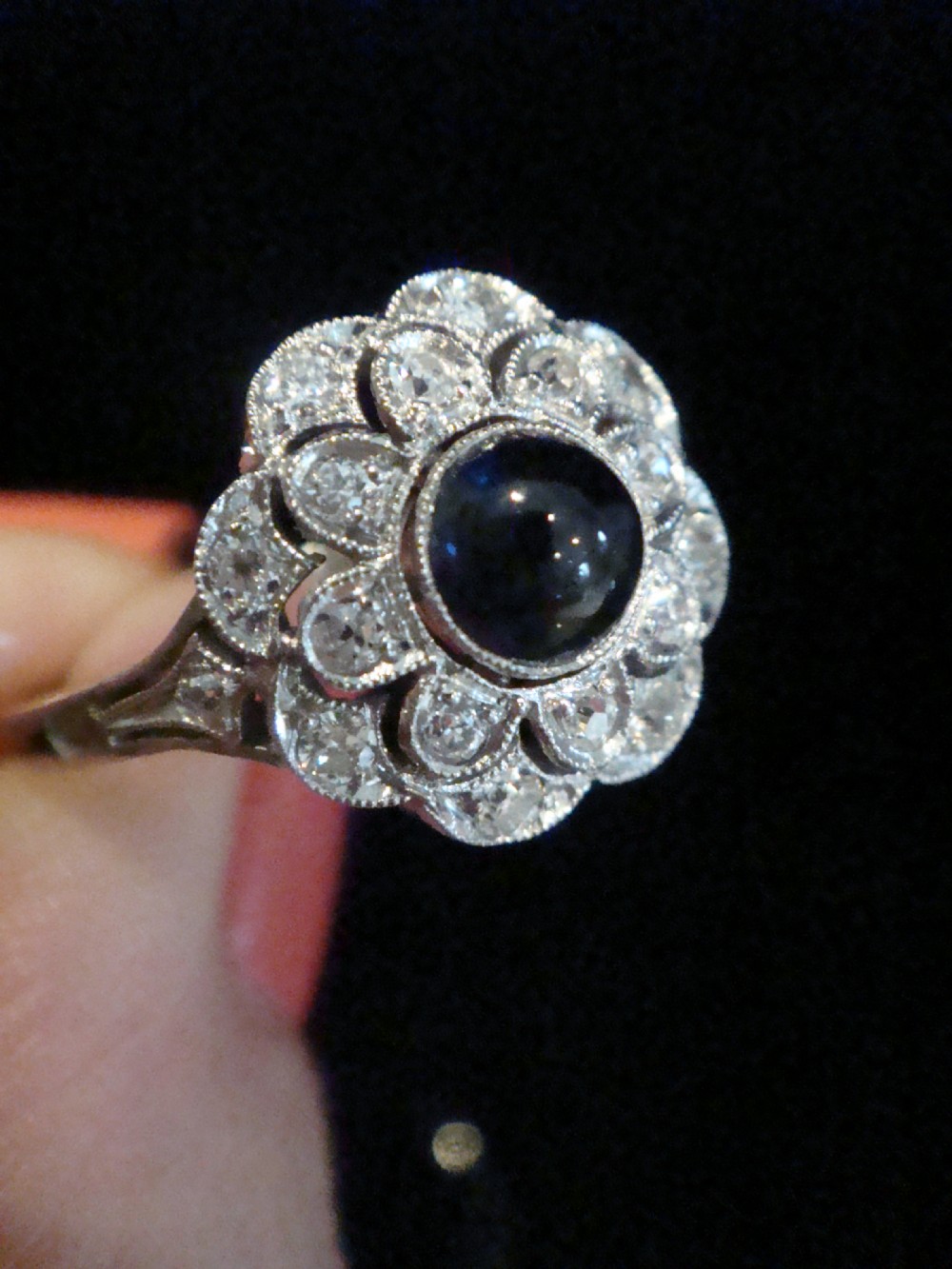 An Art Deco 18ct white gold cluster ring with a central dark blue cabochon sapphire surrounded by double scalloped halos of old cut diamonds. View VI