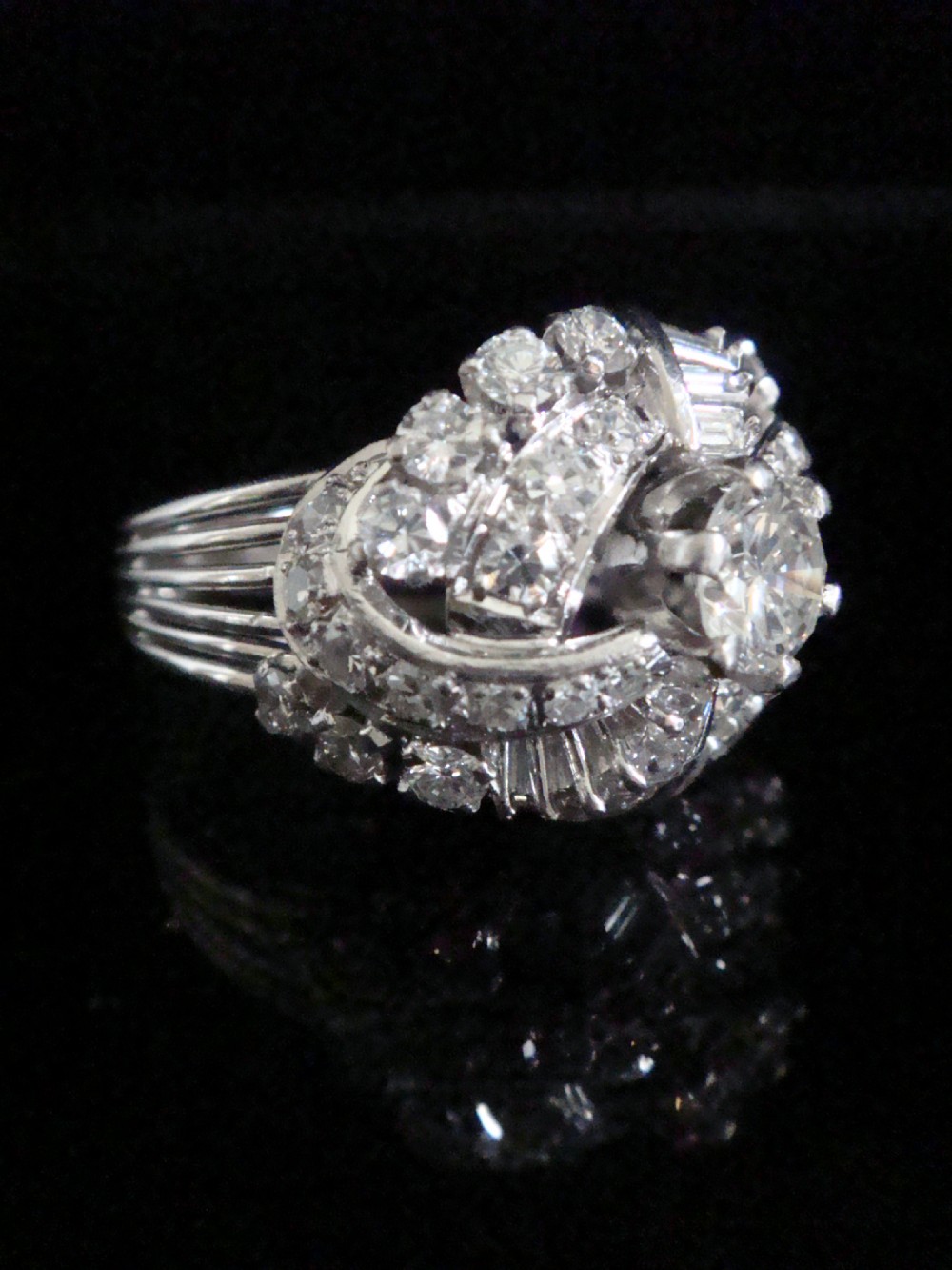 An Art Deco diamond bombe ring featuring a central transitional cut diamond surrounded by sculpted rows of round and baguette cut diamonds set in white gold. View II