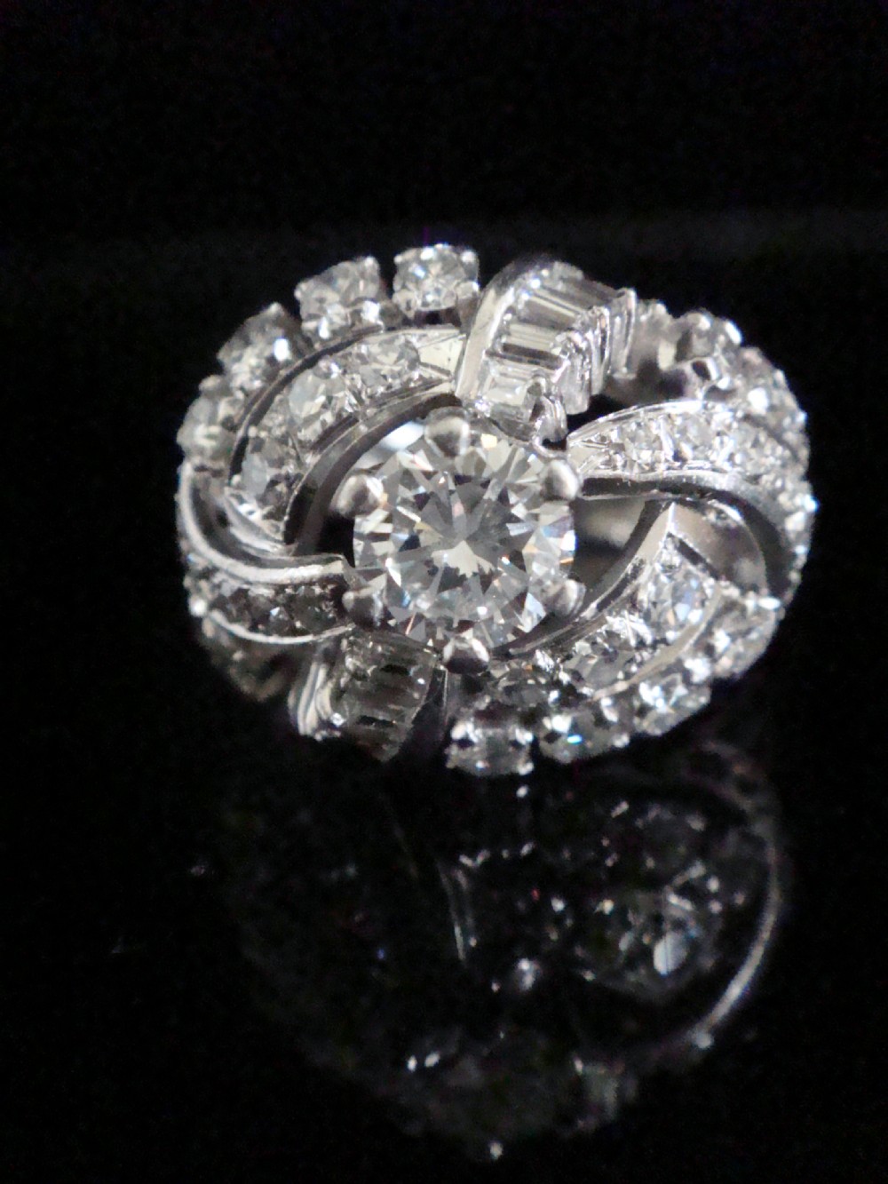 An Art Deco diamond bombe ring featuring a central transitional cut diamond surrounded by sculpted rows of round and baguette cut diamonds set in white gold.