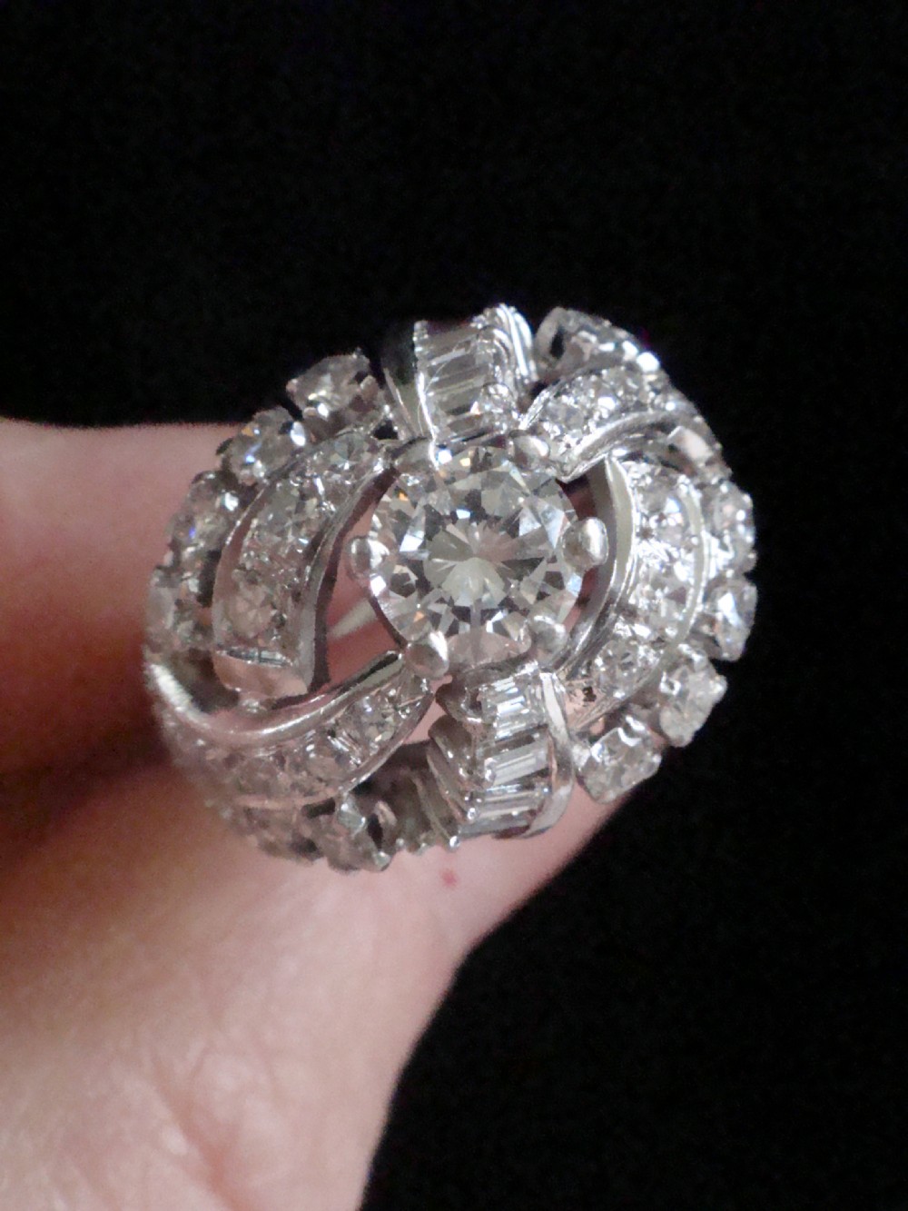 An Art Deco diamond bombe ring featuring a central transitional cut diamond surrounded by sculpted rows of round and baguette cut diamonds set in white gold. View IV
