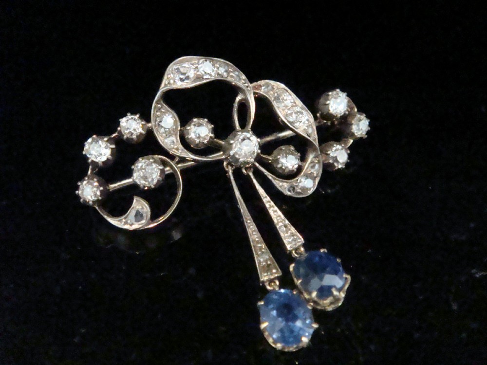 An Edwardian silver and gold bow brooch set with round diamonds and two hanging oval blue sapphires.