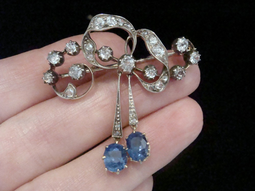 An Edwardian silver and gold bow brooch set with round diamonds and two hanging oval blue sapphires. View IV