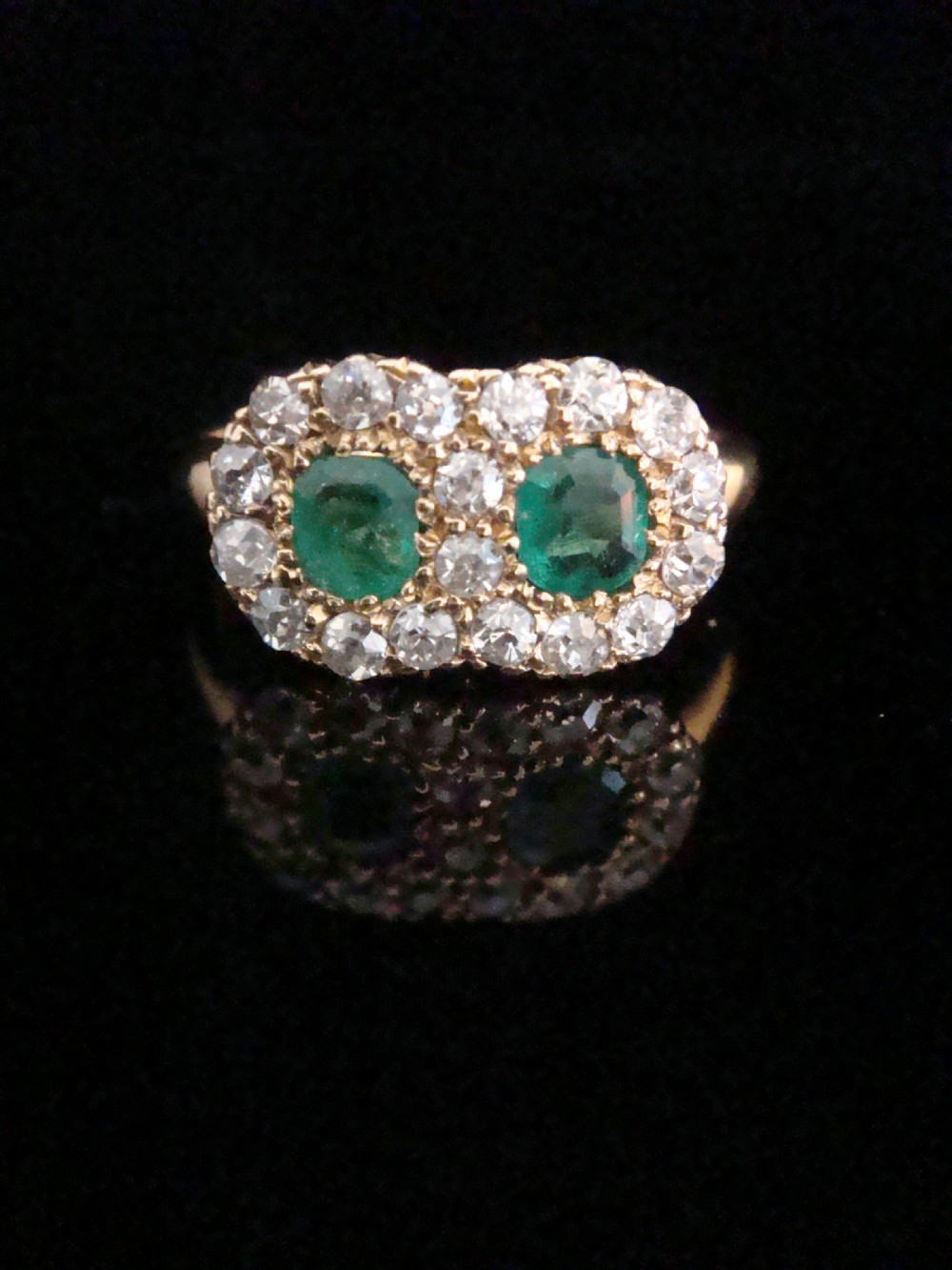 An Edwardian 18ct yellow gold ring featuring two central oval emeralds surrounded by a double cluster halo of diamonds.