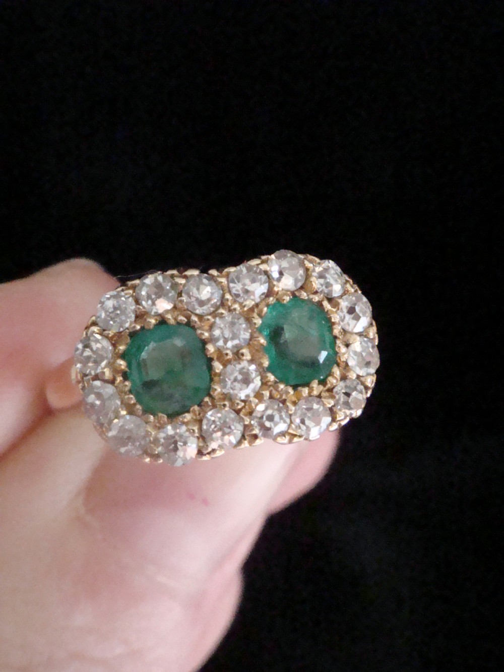 An Edwardian 18ct yellow gold ring featuring two central oval emeralds surrounded by a double cluster halo of diamonds. View IV