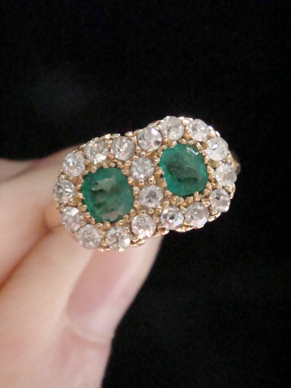 An Edwardian 18ct yellow gold ring featuring two central oval emeralds surrounded by a double cluster halo of diamonds. View V