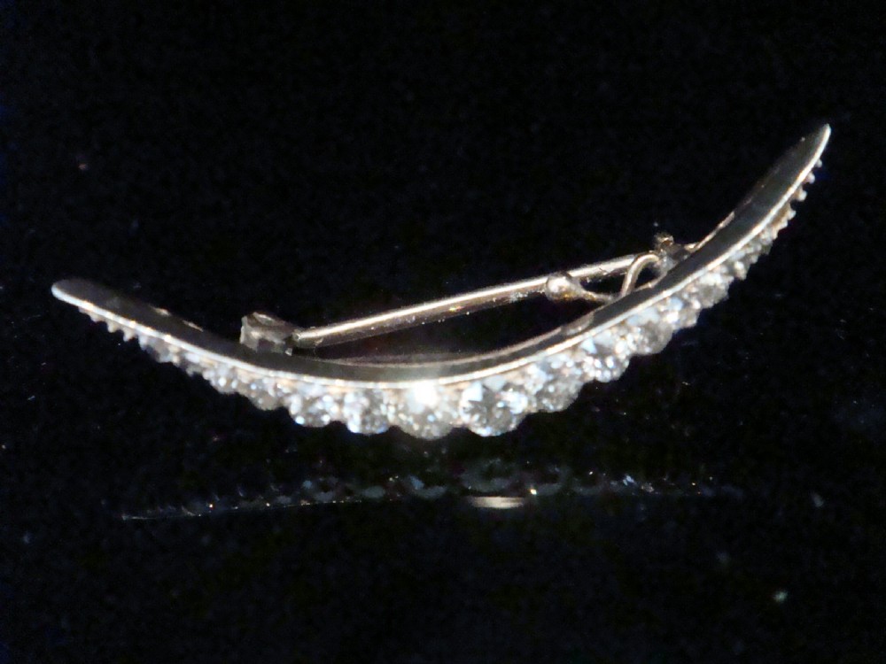 An Edwardian crescent moon brooch featuring a graduated row of round diamonds mounted in a curved precious metal setting with a pin back.