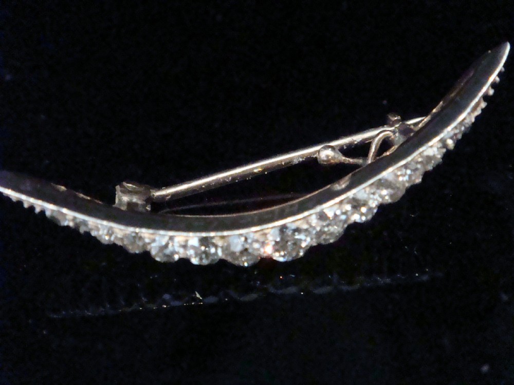 An Edwardian crescent moon brooch featuring a graduated row of round diamonds mounted in a curved precious metal setting with a pin back. View II