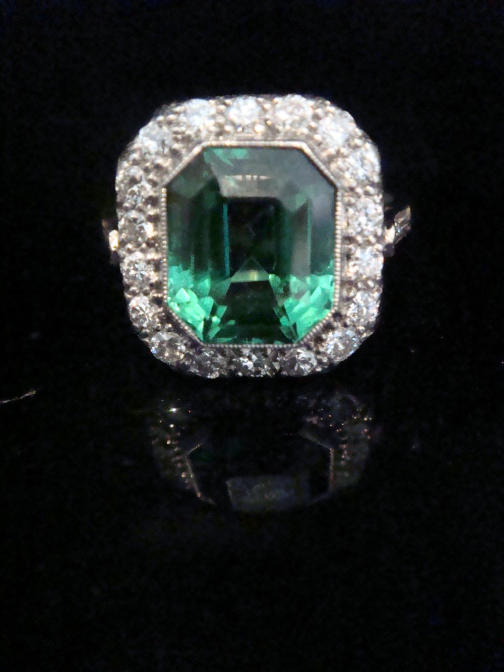 An Art Deco cluster ring featuring a central emerald-cut green tourmaline surrounded by a halo of diamonds set in platinum.