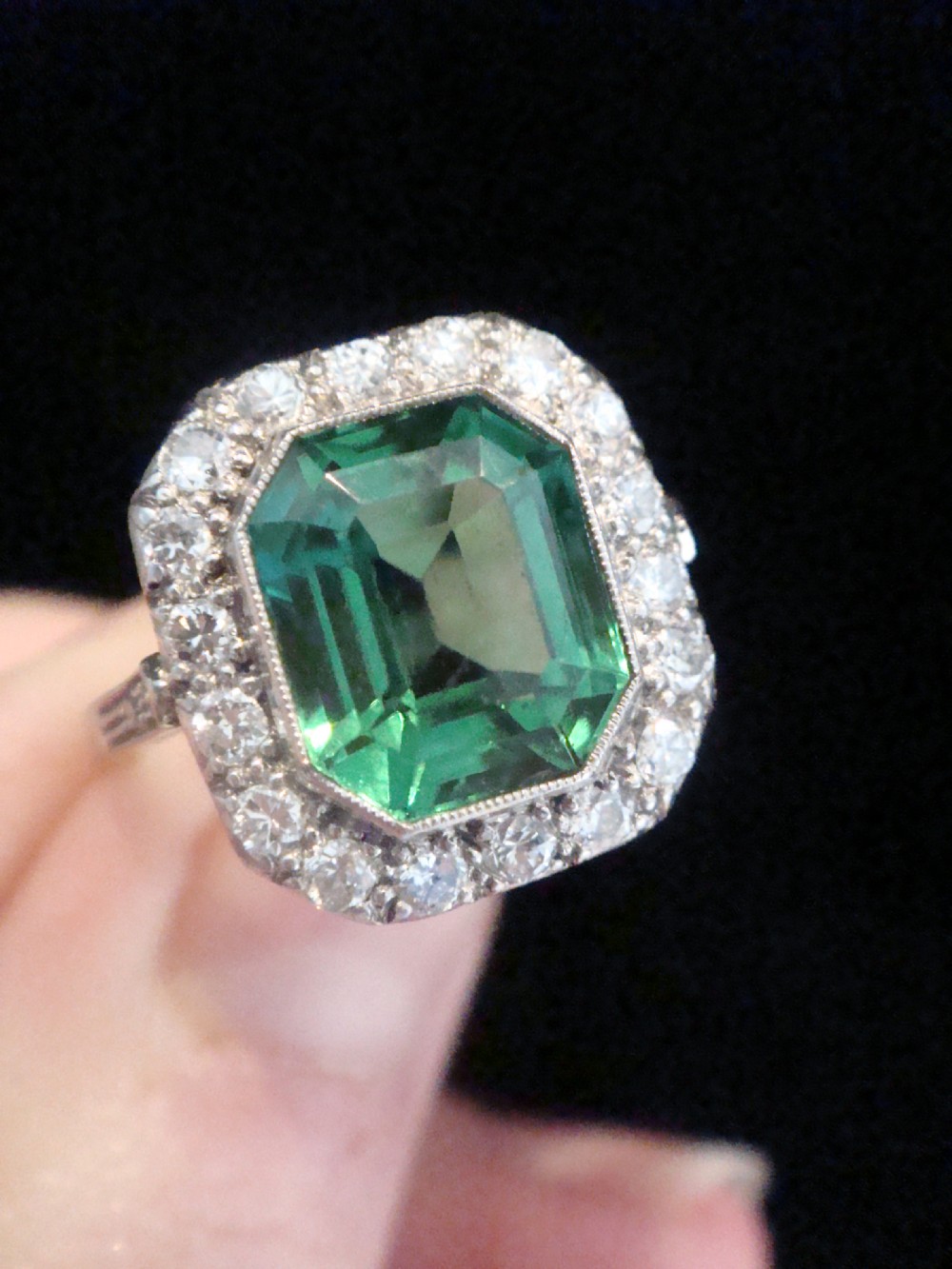 An Art Deco cluster ring featuring a central emerald-cut green tourmaline surrounded by a halo of diamonds set in platinum. View V