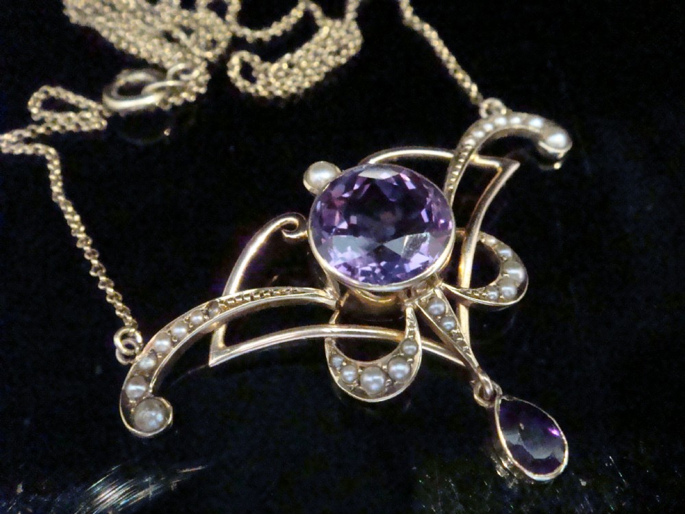 An Edwardian Art Nouveau 15ct gold lavaliere necklace featuring an openwork frame set with a round central amethyst, seed pearls, and a pear-cut dangling amethyst drop.