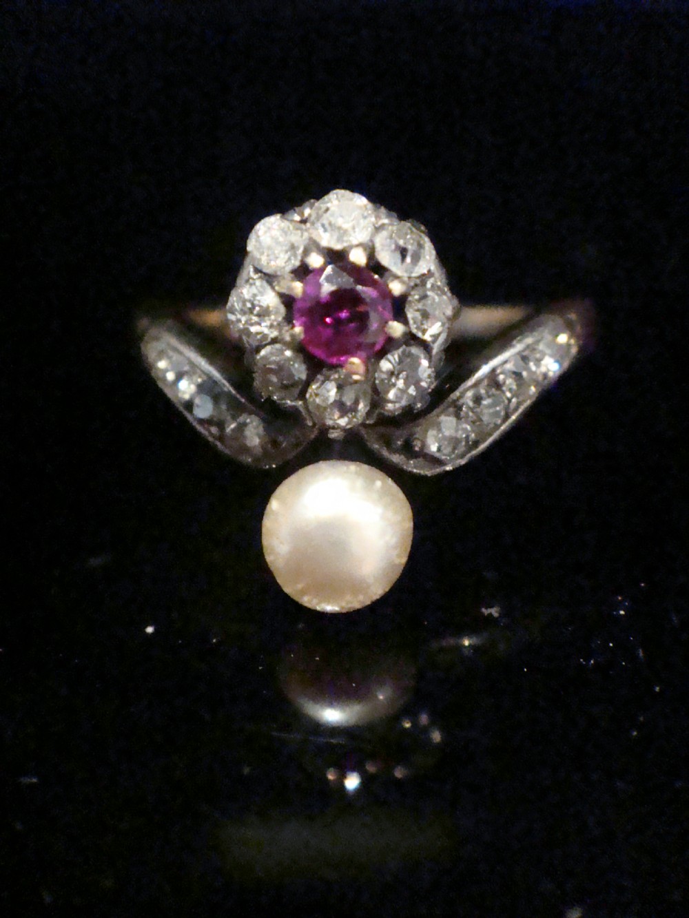 An Edwardian 18ct yellow gold ring featuring a central round ruby surrounded by old-cut diamonds with accent shoulder diamonds and a natural pearl.