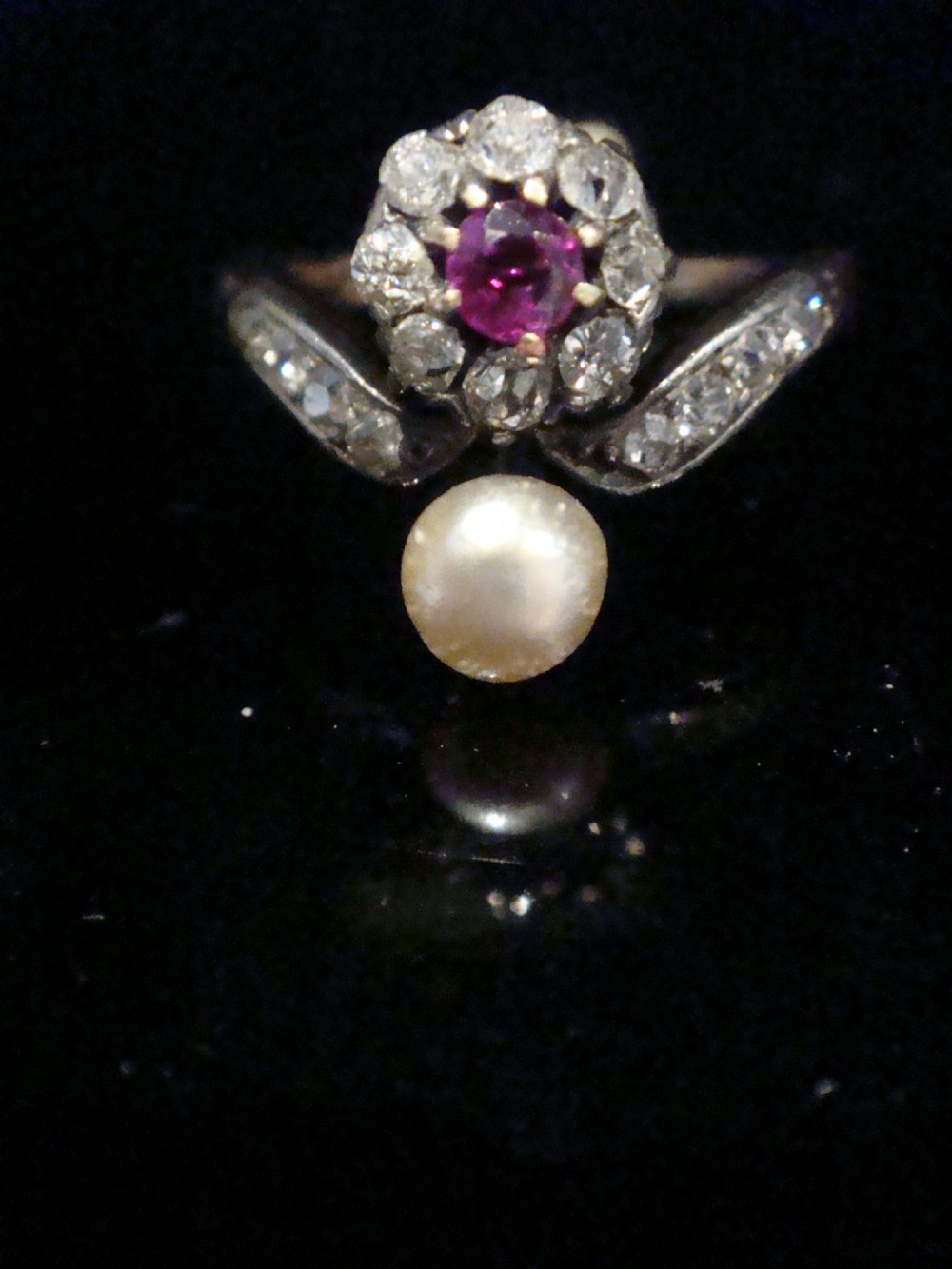 An Edwardian 18ct yellow gold ring featuring a central round ruby surrounded by old-cut diamonds with accent shoulder diamonds and a natural pearl. View V