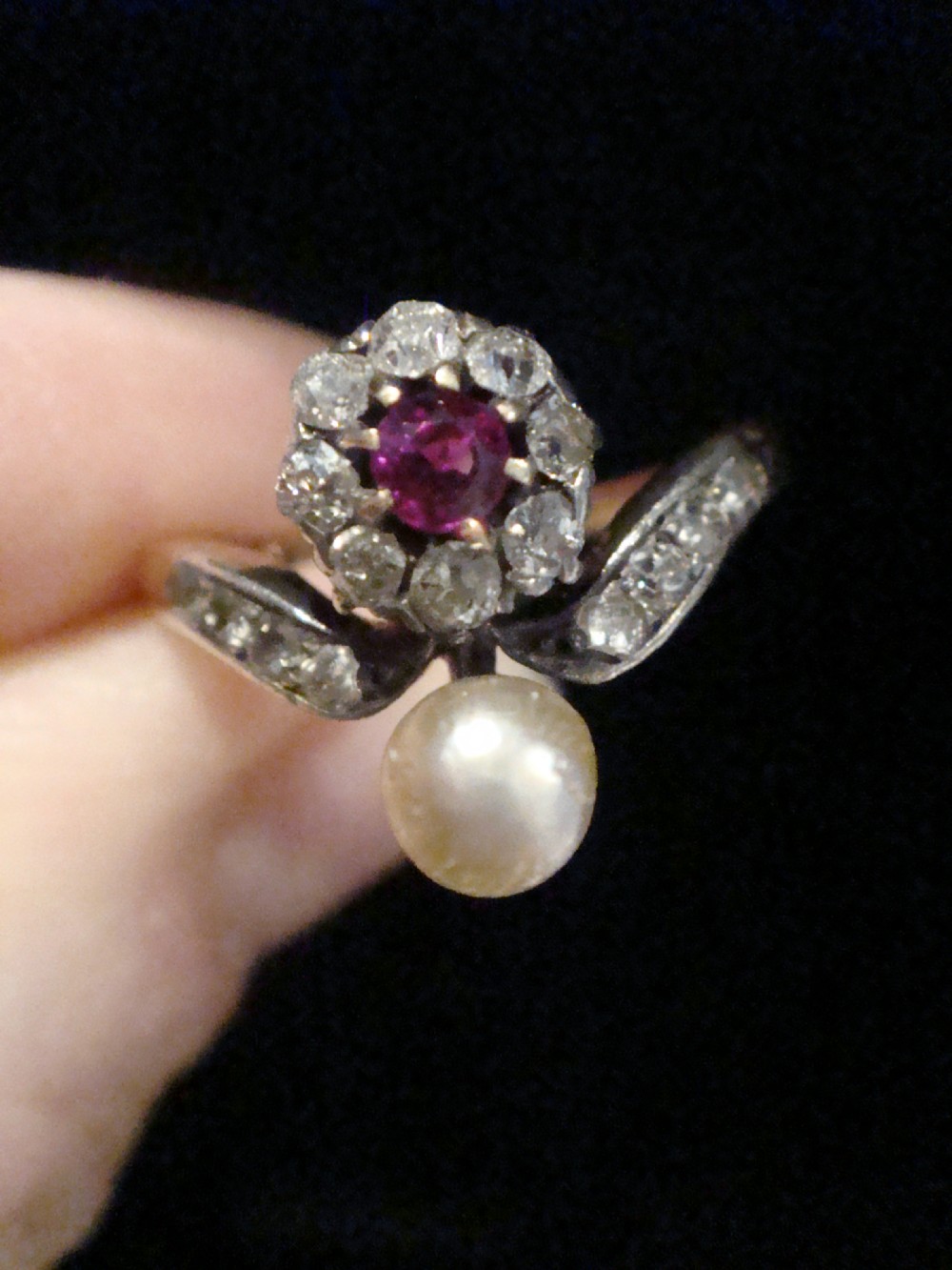 An Edwardian 18ct yellow gold ring featuring a central round ruby surrounded by old-cut diamonds with accent shoulder diamonds and a natural pearl. View VI