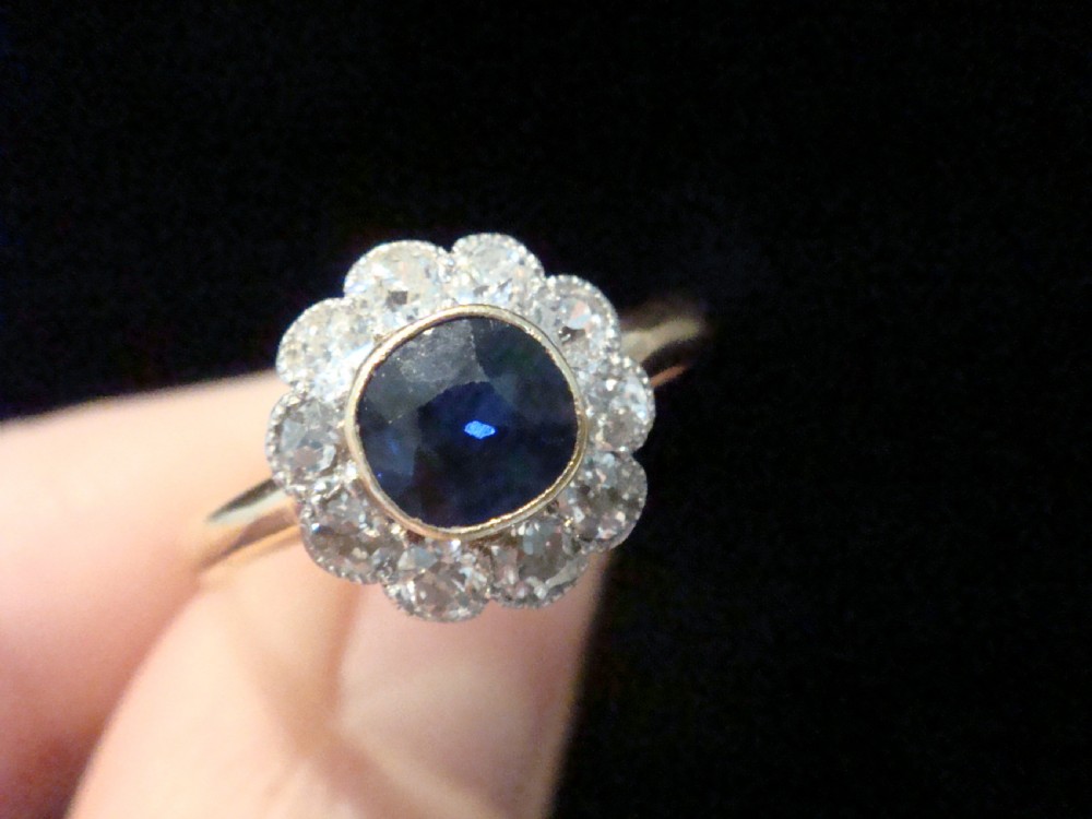 An Edwardian 18ct gold cluster ring featuring a central oval blue sapphire surrounded by a scalloped halo of small diamonds.