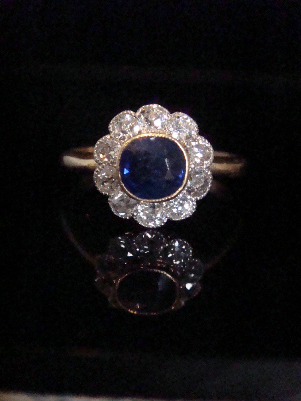 An Edwardian 18ct gold cluster ring featuring a central oval blue sapphire surrounded by a scalloped halo of small diamonds. View II