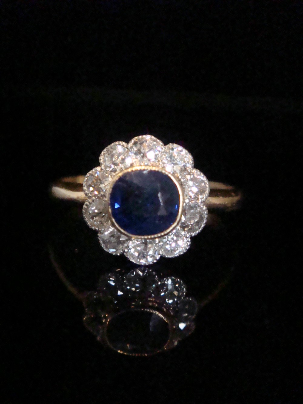 An Edwardian 18ct gold cluster ring featuring a central oval blue sapphire surrounded by a scalloped halo of small diamonds. View III