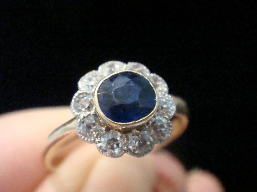 An Edwardian 18ct gold cluster ring featuring a central oval blue sapphire surrounded by a scalloped halo of small diamonds. View VI