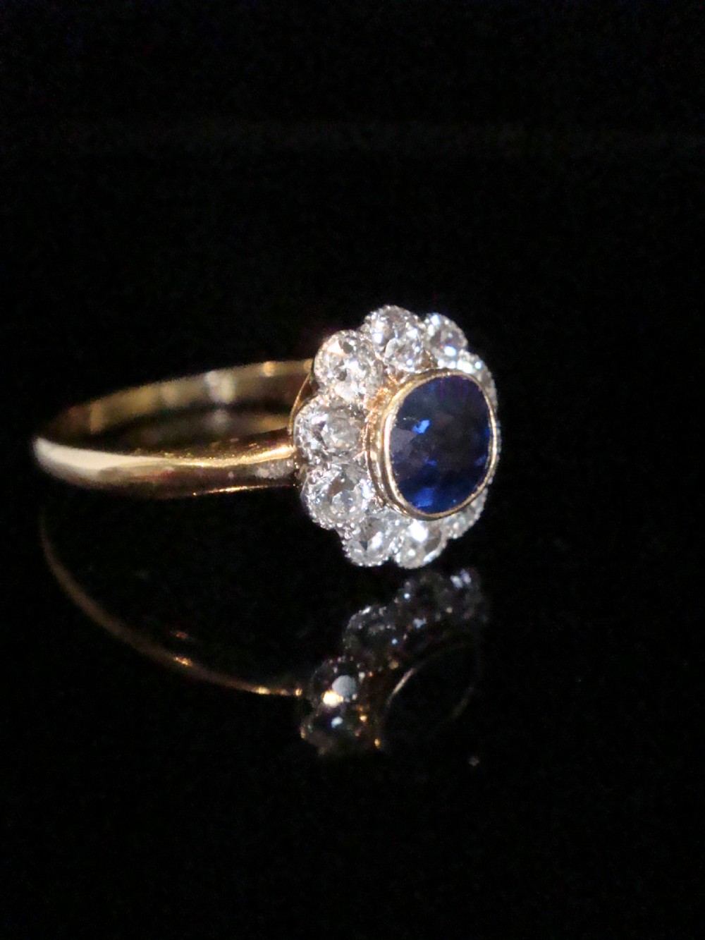 An Edwardian 18ct gold cluster ring featuring a central oval blue sapphire surrounded by a scalloped halo of small diamonds. View VII