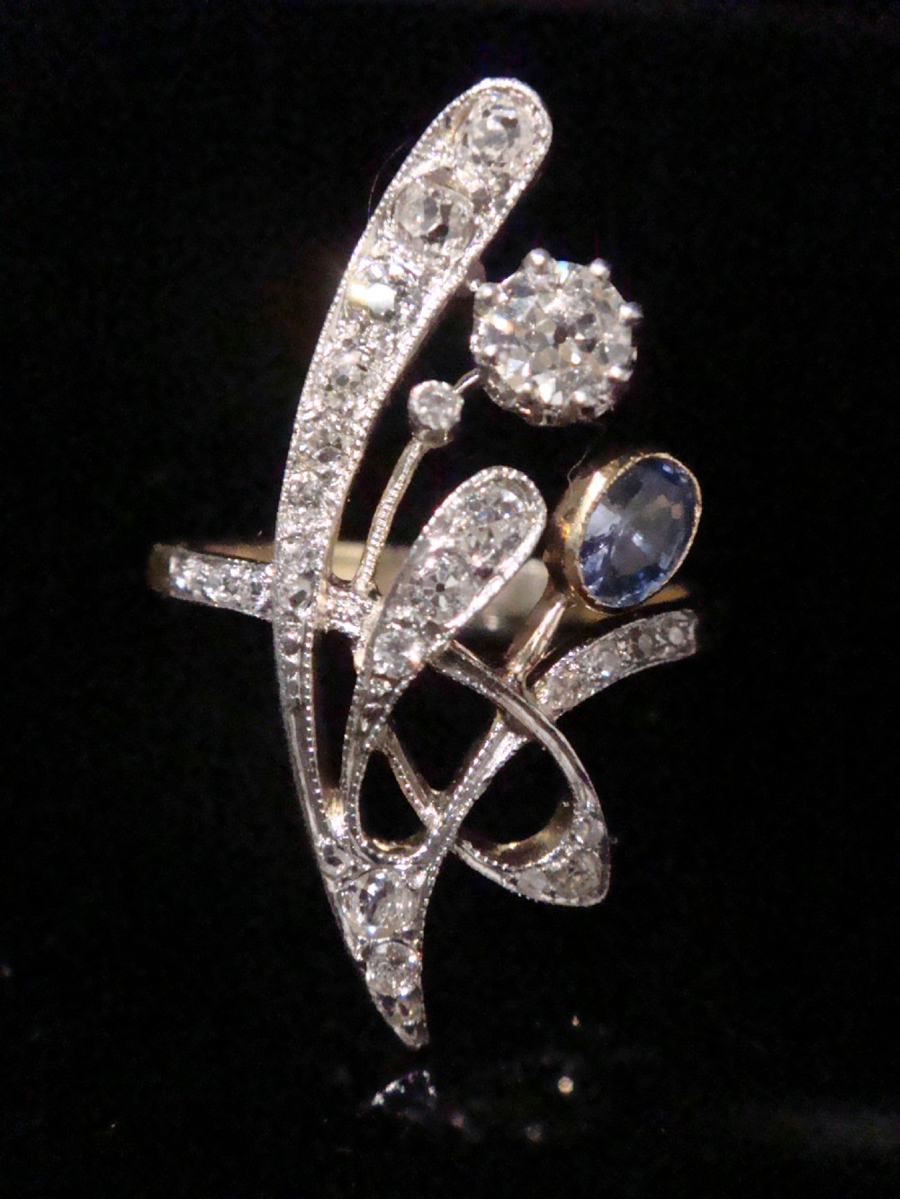 An Art Nouveau asymmetrical ring featuring fluid platinum bands encrusted with diamonds leading to a central diamond and an oval blue sapphire set in gold.
