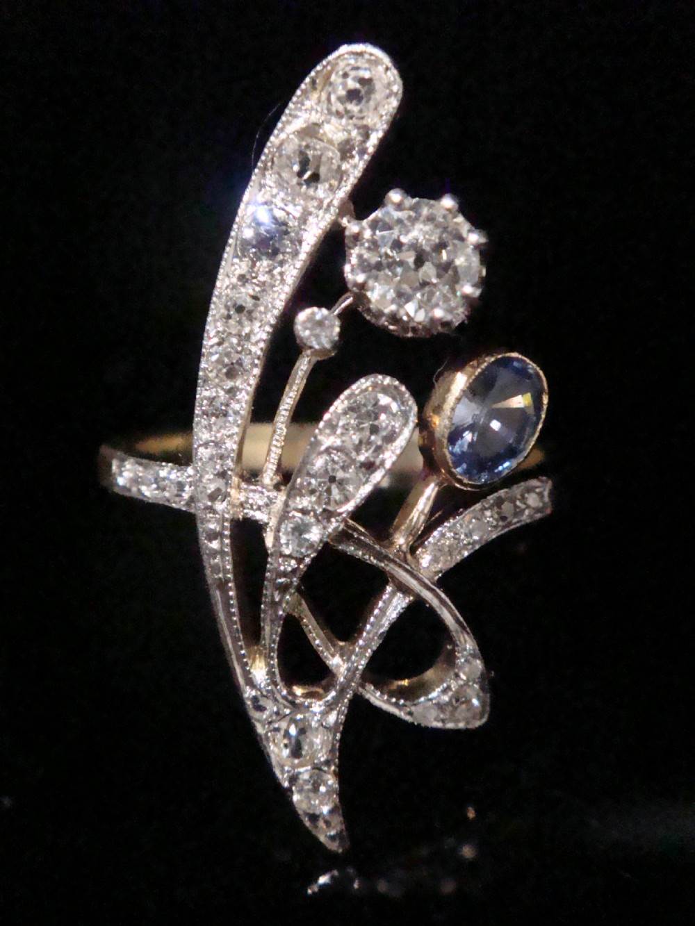 An Art Nouveau asymmetrical ring featuring fluid platinum bands encrusted with diamonds leading to a central diamond and an oval blue sapphire set in gold. View IV