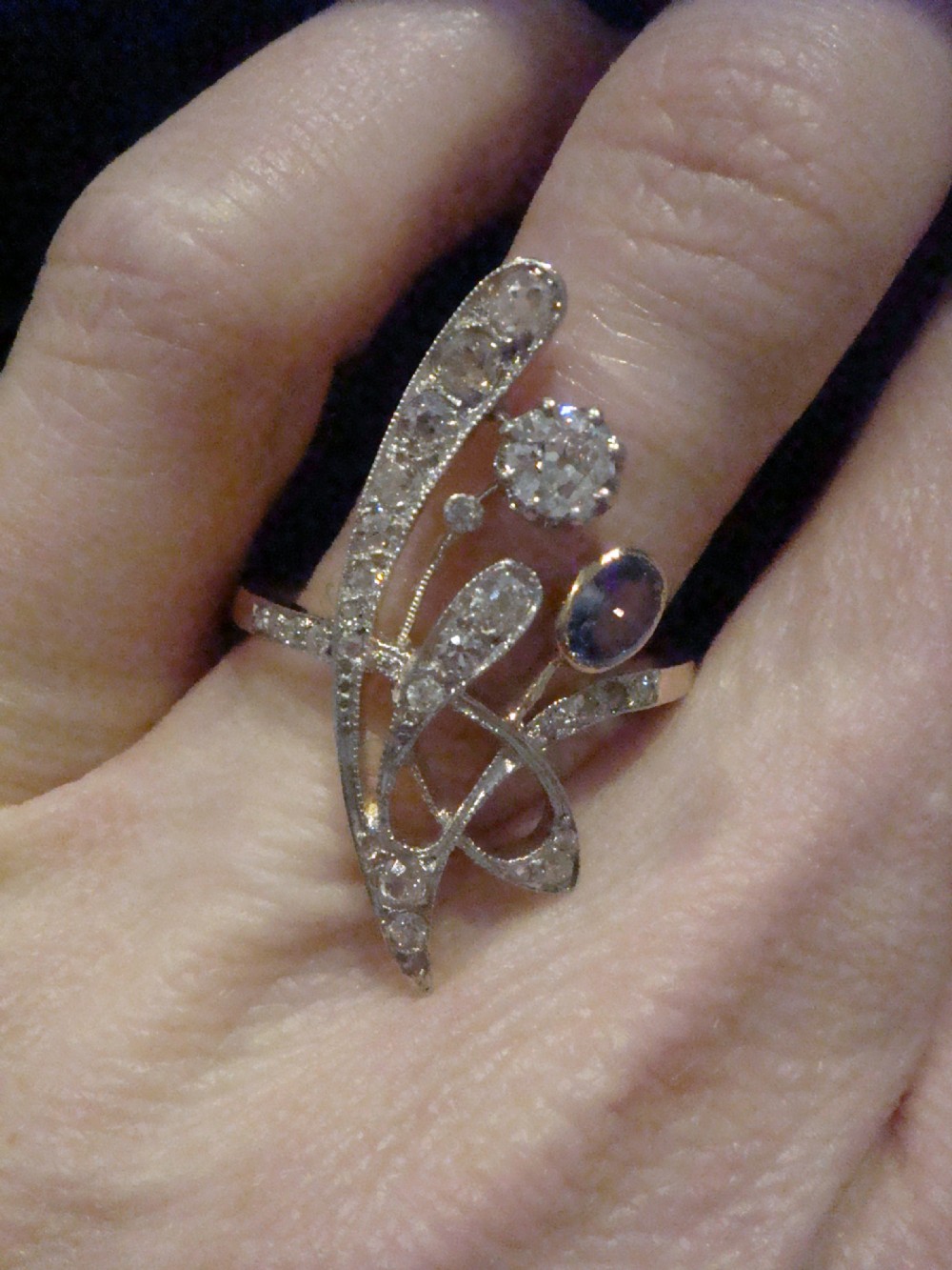 An Art Nouveau asymmetrical ring featuring fluid platinum bands encrusted with diamonds leading to a central diamond and an oval blue sapphire set in gold. View V