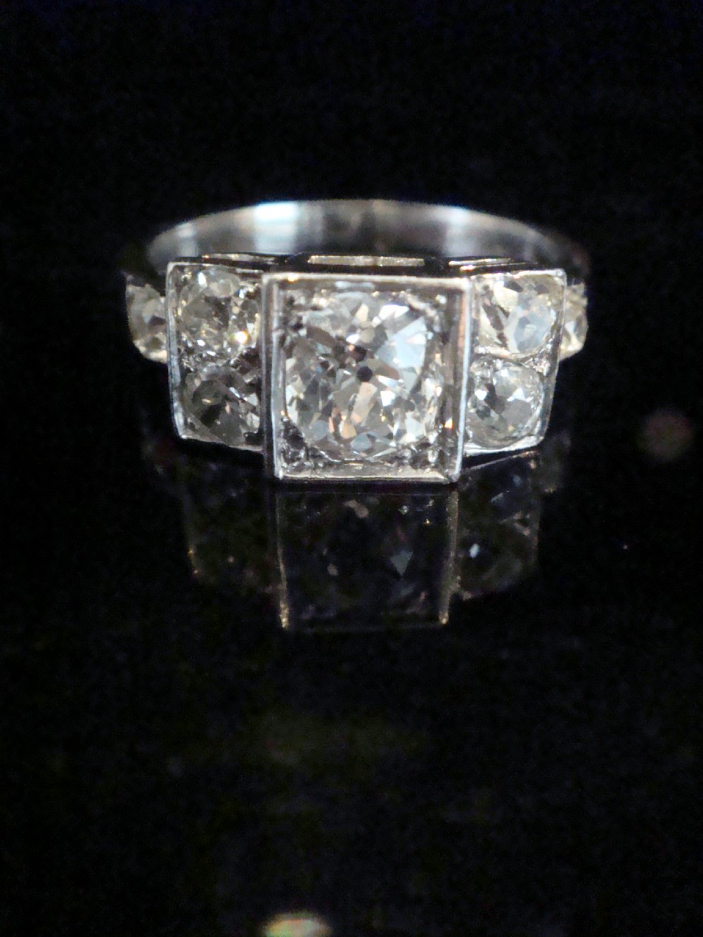 An Art Deco 18ct white gold ring featuring a central round old cut diamond flanked by smaller diamonds set within a geometric square panel.