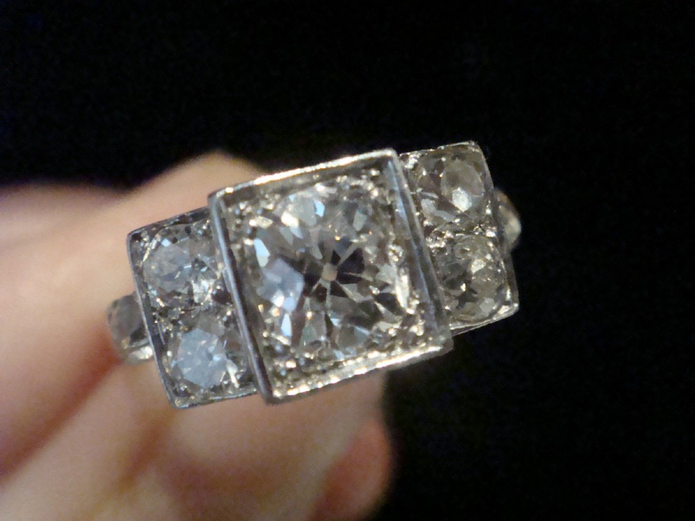 An Art Deco 18ct white gold ring featuring a central round old cut diamond flanked by smaller diamonds set within a geometric square panel. View II