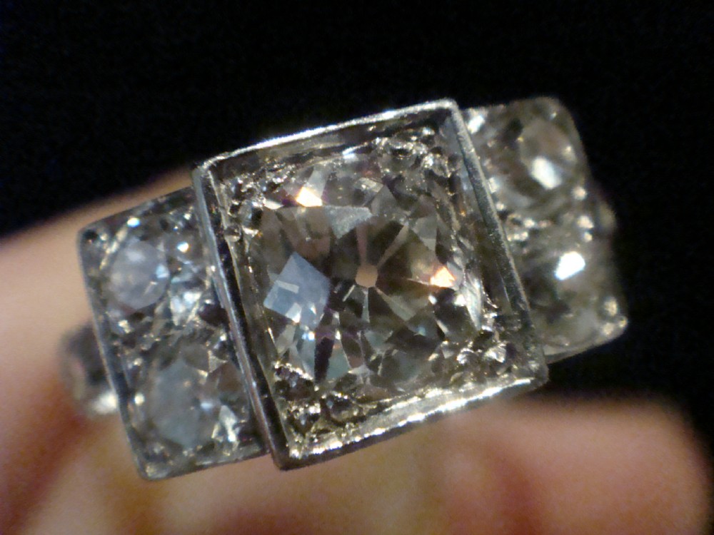 An Art Deco 18ct white gold ring featuring a central round old cut diamond flanked by smaller diamonds set within a geometric square panel. View III