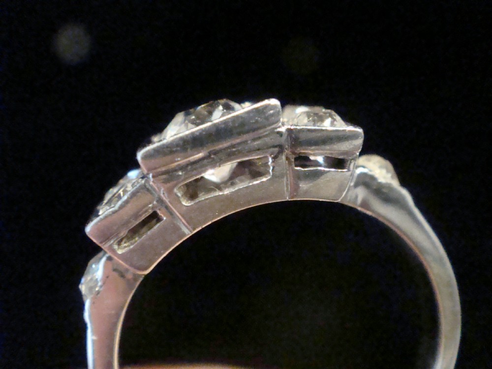 An Art Deco 18ct white gold ring featuring a central round old cut diamond flanked by smaller diamonds set within a geometric square panel. View IV