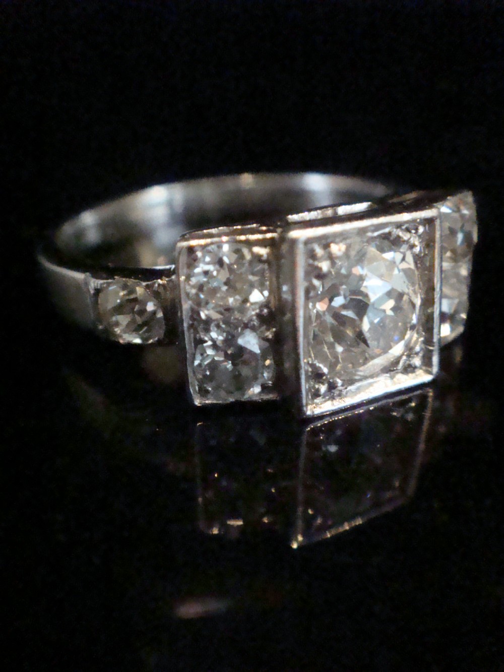 An Art Deco 18ct white gold ring featuring a central round old cut diamond flanked by smaller diamonds set within a geometric square panel. View V