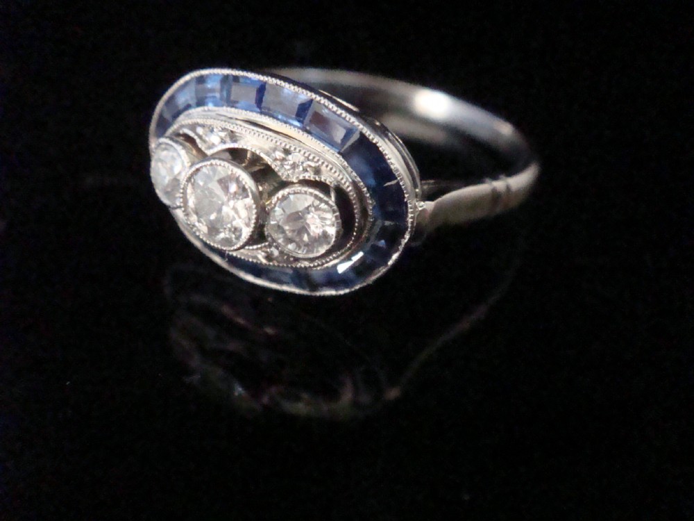 An Art Deco 18ct white gold ring featuring three central diamonds framed by milgrain details and an outer oval border of calibre-cut blue sapphires.