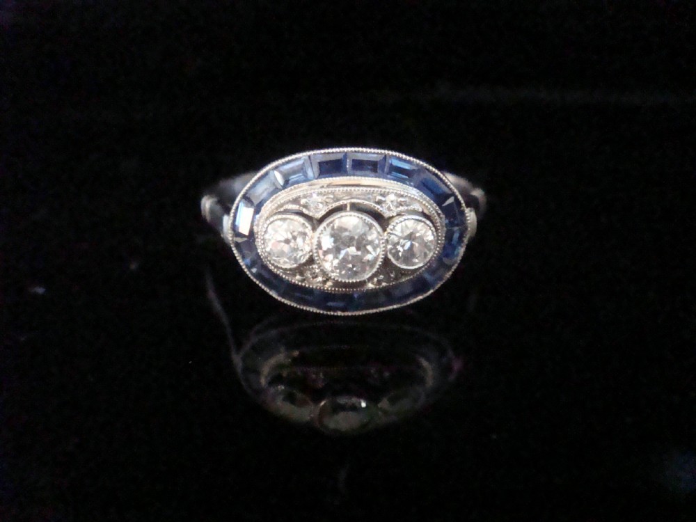 An Art Deco 18ct white gold ring featuring three central diamonds framed by milgrain details and an outer oval border of calibre-cut blue sapphires. View II